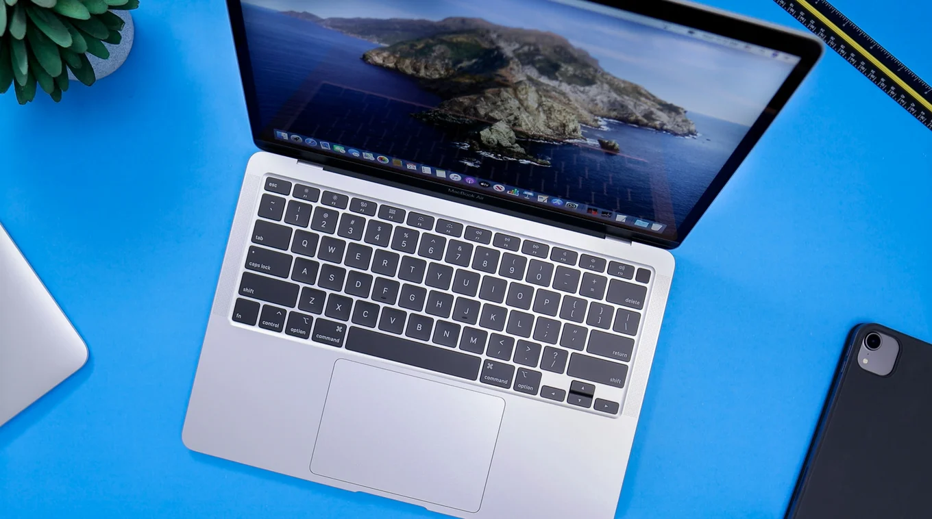 A top-down view of a macbook computer laptop with keyboard on a blue background. A black iphone, green succulent, black and yellow ruler, and silver corner of another device are also in the frame.